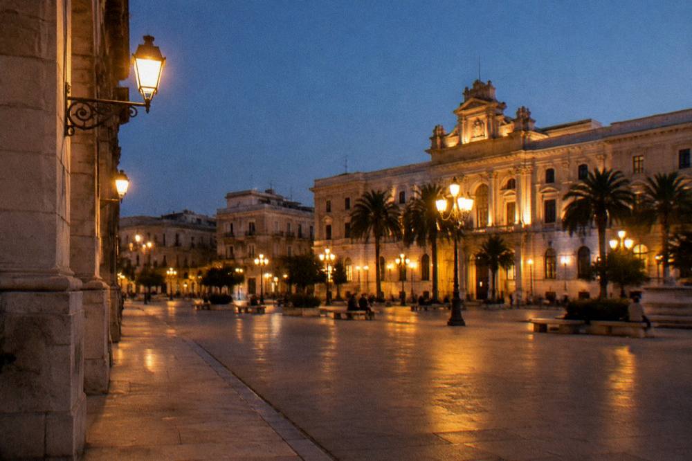 City square at evening with lit street lamps, distant people on benches and a warmly lit stone facade