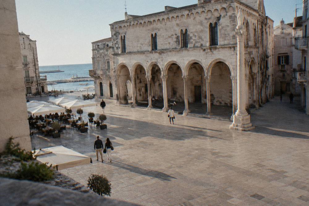 Old town square with a medieval arcaded loggia, a slender stone column and cafe tables under parasols in soft morning light