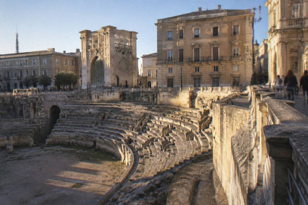Roman amphitheatre half excavated in a city square surrounded by palazzi in afternoon light