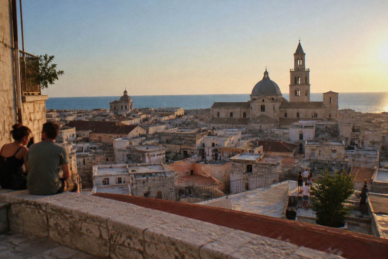 Terracotta and limestone rooftops and church domes of a dense old town seen from a high terrace with the sea beyond