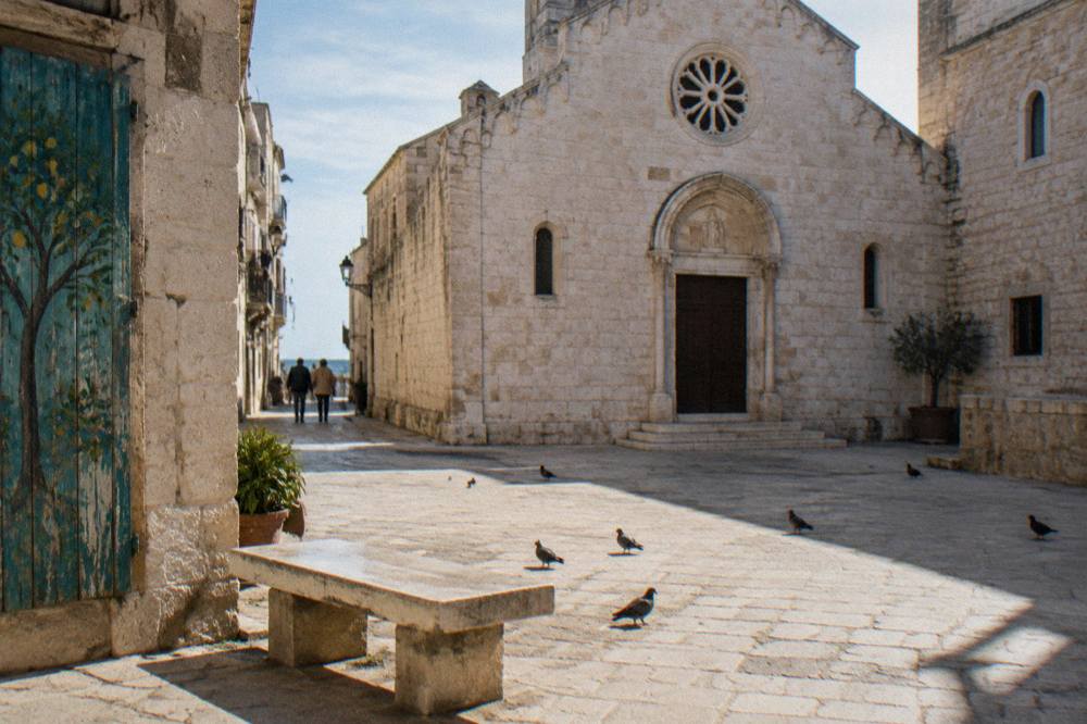 Small stone piazza with a modest pale limestone church front, a stone bench and pigeons in strong midday light