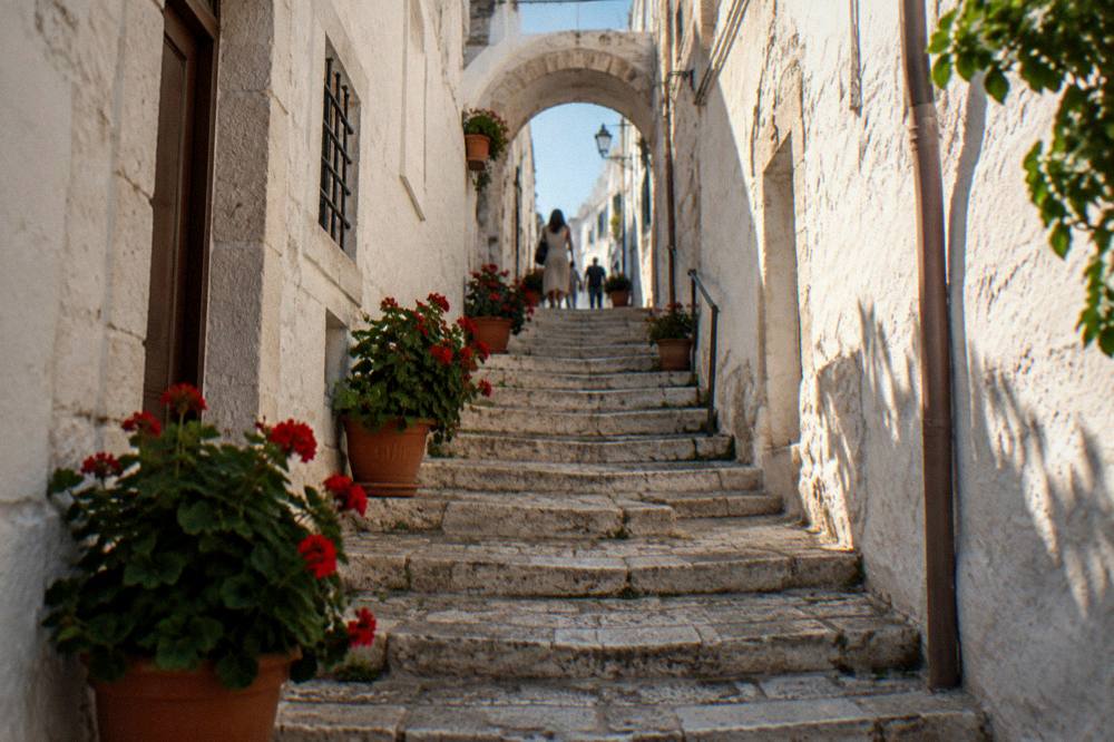 Narrow lane with a steep stone staircase climbing between whitewashed walls to a small arch with potted geraniums