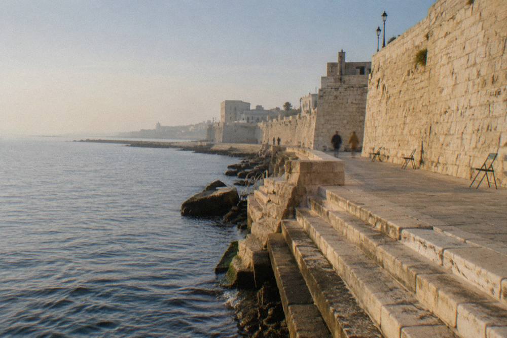 Massive pale limestone sea wall with a promenade at its foot and worn steps down to the water in low afternoon sun