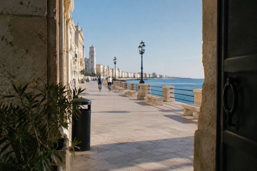 Cyclists far off passing stone benches on a promenade between cream monumental buildings and the calm blue sea