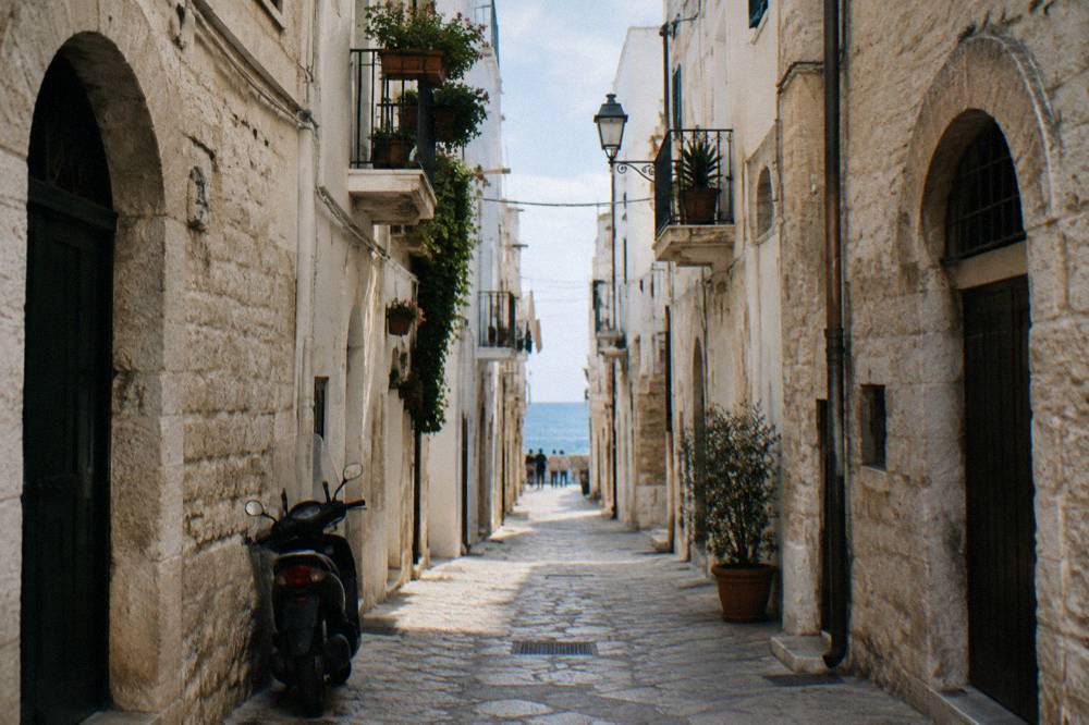 Straight old town lane between whitewashed walls ending in a bright slit of open sea with a parked scooter