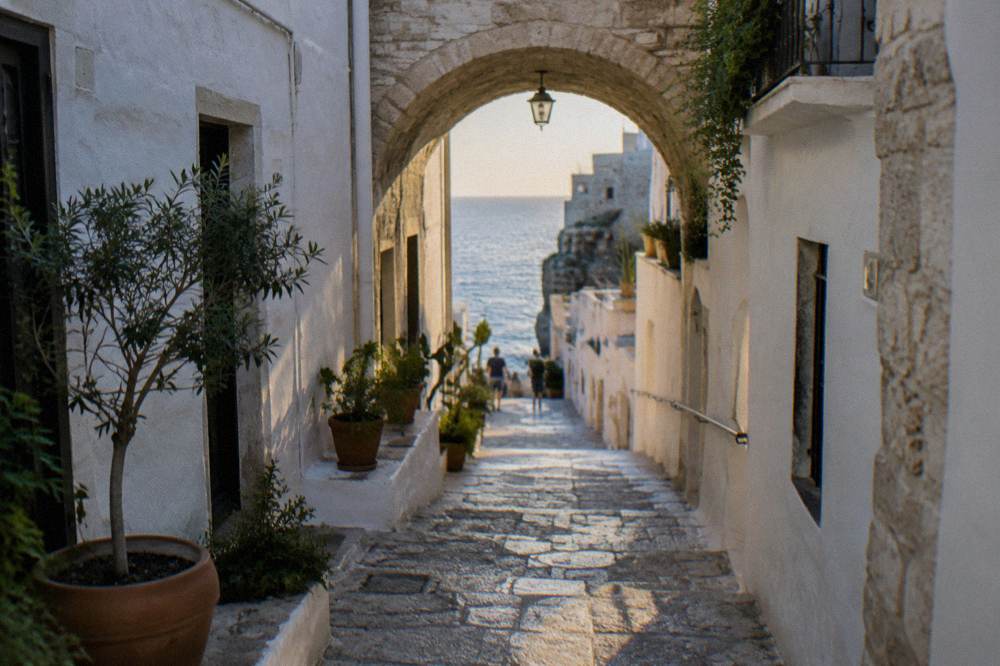 Narrow whitewashed cliff-top old town lane with a stone arch, potted plants and a glimpse of blue sea