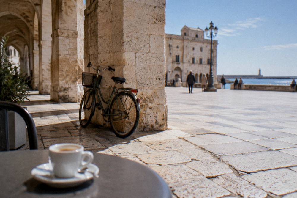 Worn limestone paving beside loggia arches with a leaning bicycle and an espresso cup on a small round table