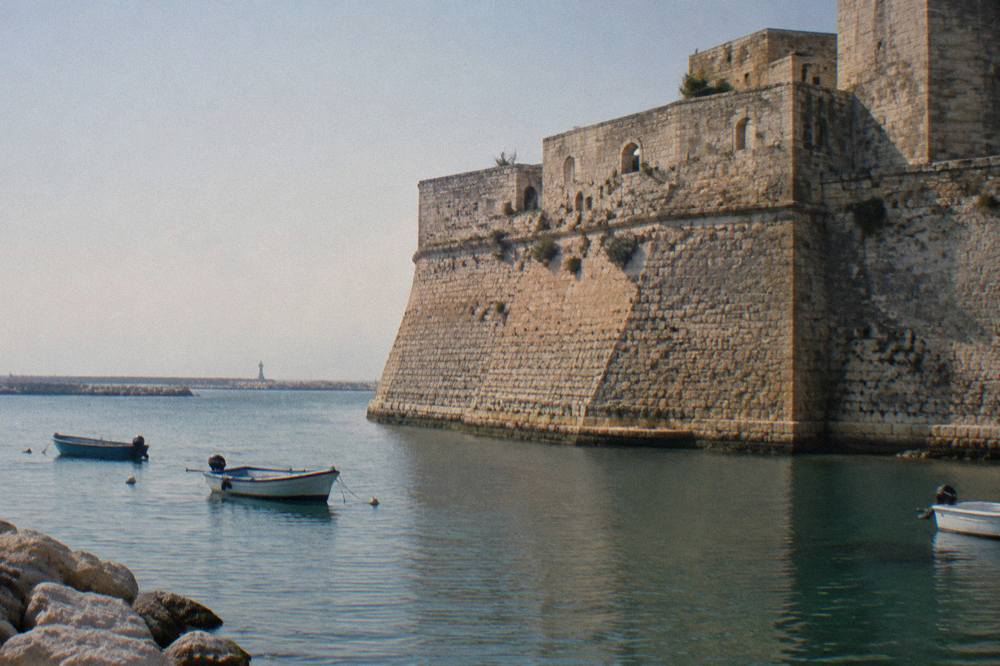 Seaward wall of a pale stone castle beside harbour water with a few small boats in afternoon sun