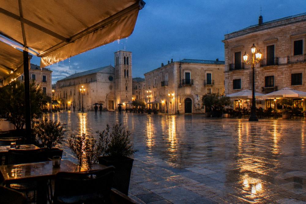 Rainy evening in a pale stone square with wet paving reflecting warm cafe lights and distant umbrellas