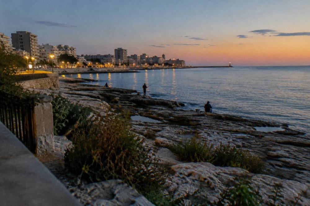 Seafront limestone rocks below a residential district with distant anglers and calm water in early evening