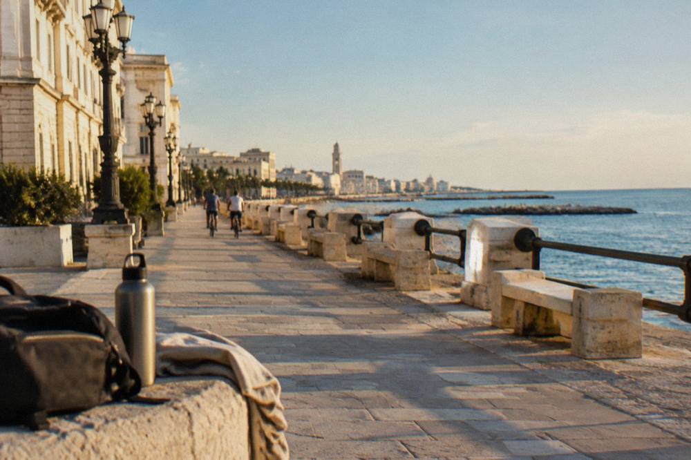 Cyclists far off passing stone benches on a promenade between cream monumental buildings and the calm blue sea