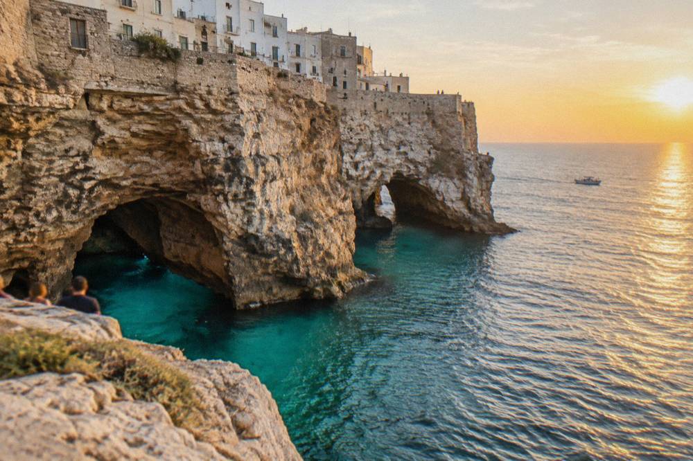 Sea cave openings at the base of tall limestone cliffs with turquoise water and a small boat far off