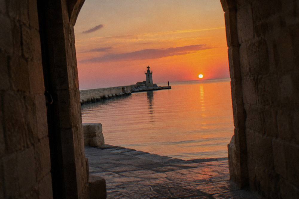 End of a harbour pier with a small lighthouse, glassy open Adriatic and a distant angler at sunset