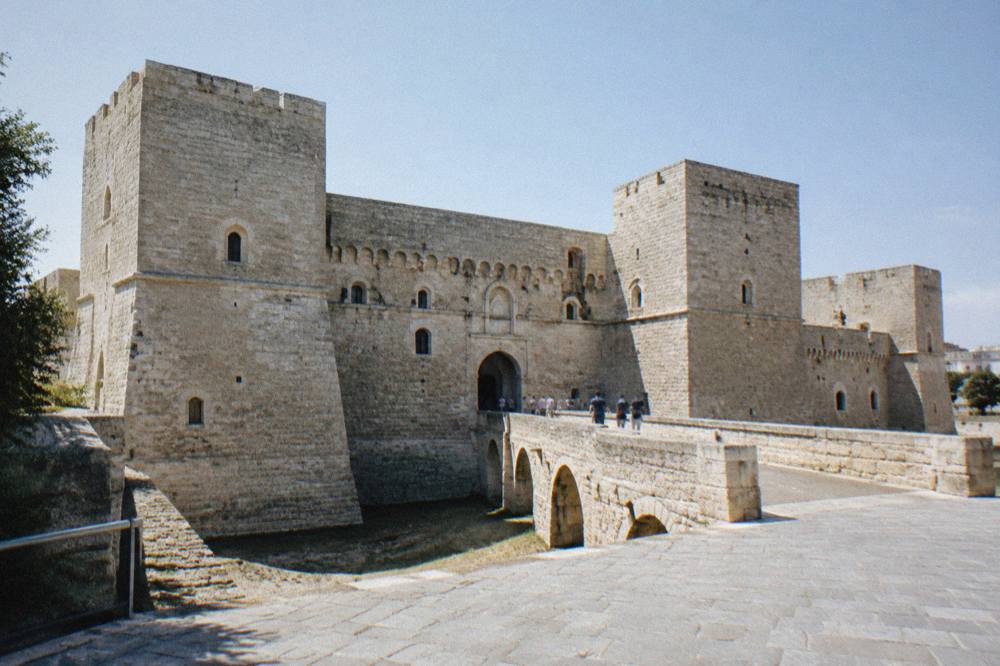 Massive square pale limestone fortress with corner towers, a dry moat and a bridge leading to an arched gate
