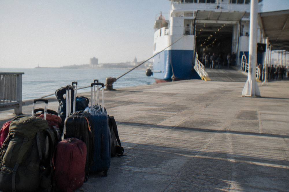 Suitcases and rucksacks stacked on a sunlit quay beside a ferry gangway with travellers waiting in the shade