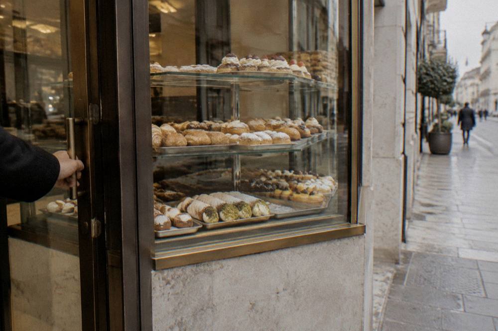 Pastry shop window with trays of pastries behind glass and a hand reaching for the door