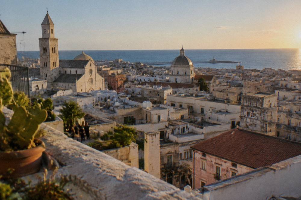 Terracotta and limestone rooftops and church domes of a dense old town seen from a high terrace with the sea beyond