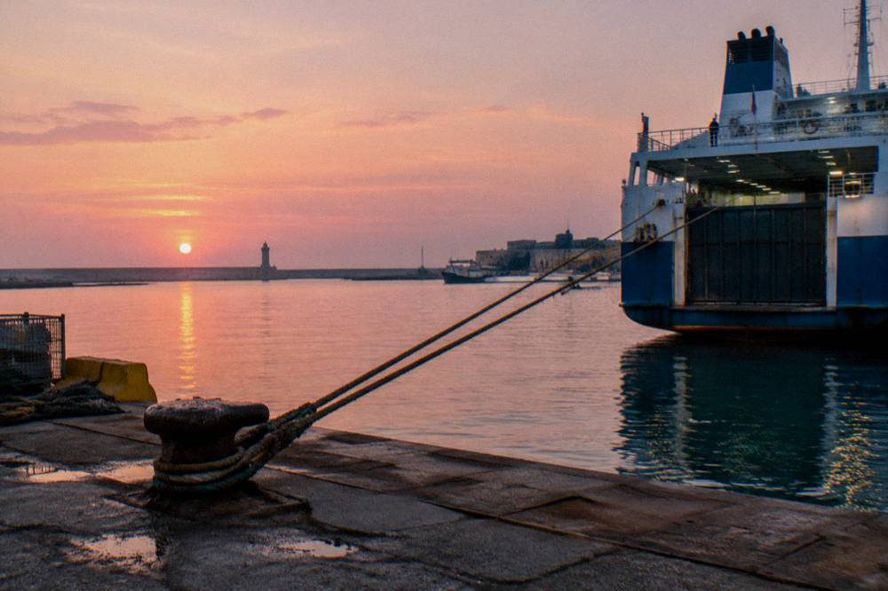 Stern of a ferry at a quay at sunrise with a pink sky, taut mooring ropes and still water