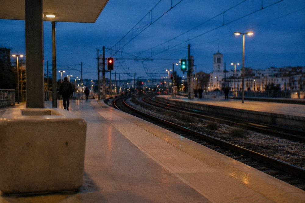 Rails curving away from a platform end at dusk with glowing signal lights and a few distant passengers