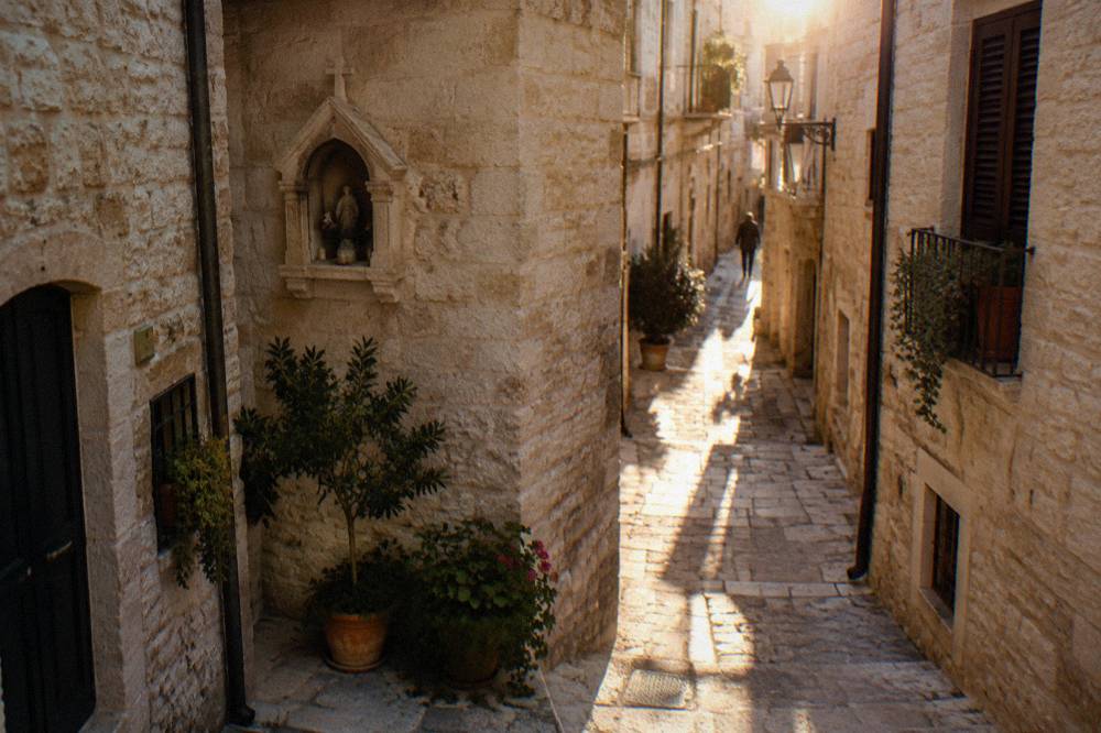 Crossing of narrow old town lanes with a stone shrine niche, potted plants and sunlight slicing between buildings