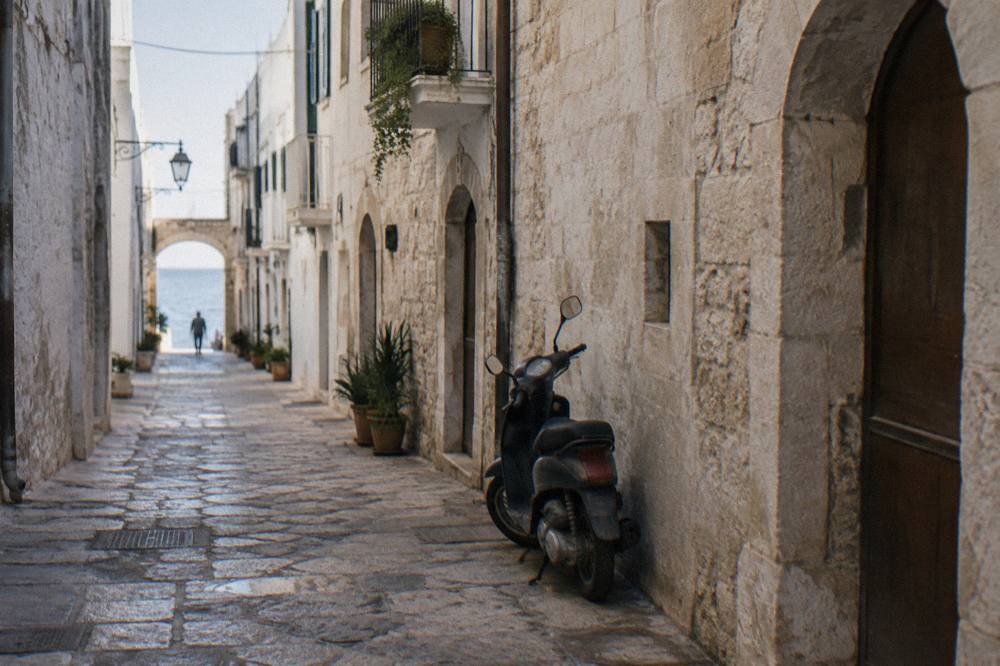 Straight old town lane between whitewashed walls ending in a bright slit of open sea with a parked scooter