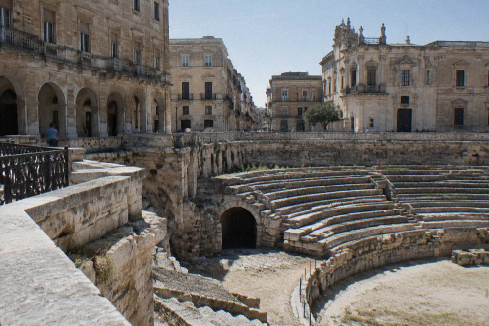 Roman amphitheatre half excavated in a city square surrounded by palazzi in afternoon light