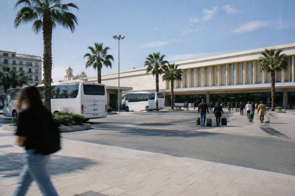 Station forecourt with plain white buses, palm trees and distant travellers with wheeled suitcases