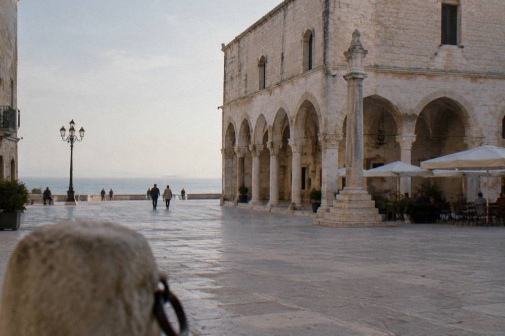 Old town square with a medieval arcaded loggia, a slender stone column and cafe tables under parasols in soft morning light