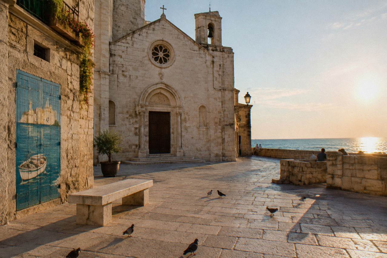Small stone piazza with a modest pale limestone church front, a stone bench and pigeons in strong midday light
