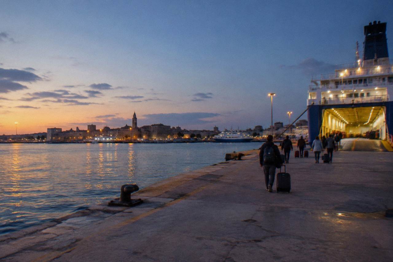 Distant passengers with wheeled suitcases walking along a quay toward a ferry's open loading ramp in evening light