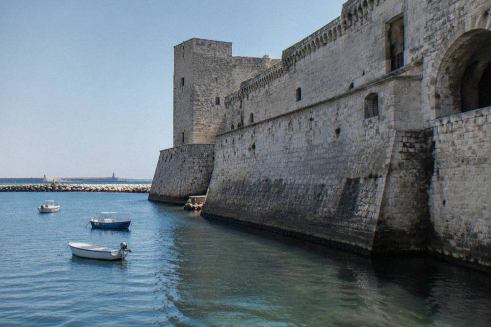 Seaward wall of a pale stone castle beside harbour water with a few small boats in afternoon sun