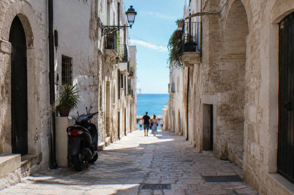 Straight old town lane between whitewashed walls ending in a bright slit of open sea with a parked scooter