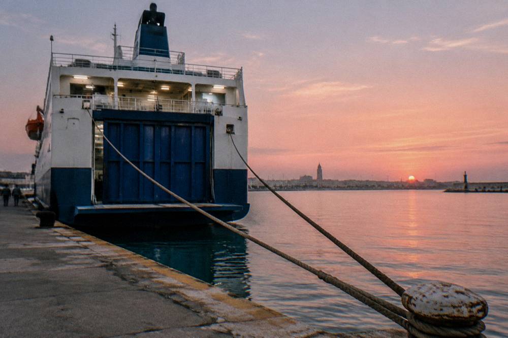 Stern of a ferry at a quay at sunrise with a pink sky, taut mooring ropes and still water