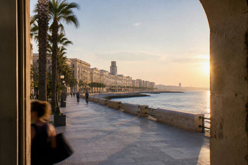 Broad seafront promenade beside a low sea wall with palm trees and a long curve of cream buildings