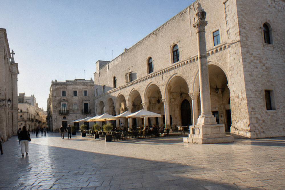 Old town square with a medieval arcaded loggia, a slender stone column and cafe tables under parasols in soft morning light