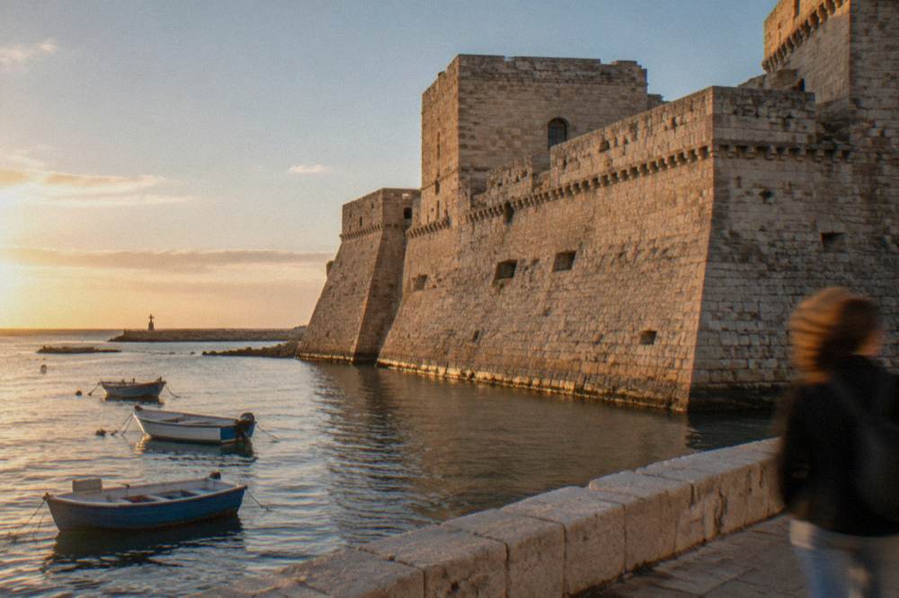 Seaward wall of a pale stone castle beside harbour water with a few small boats in afternoon sun
