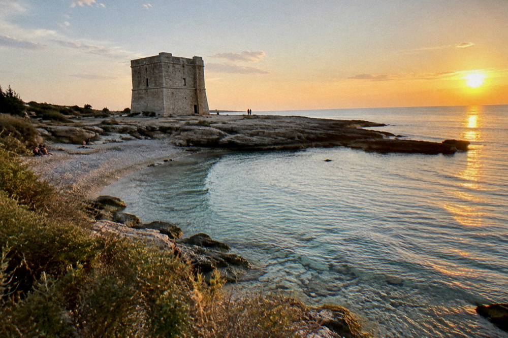 Squat square stone watchtower on a flat rocky shore beside a small pebble beach and low scrub