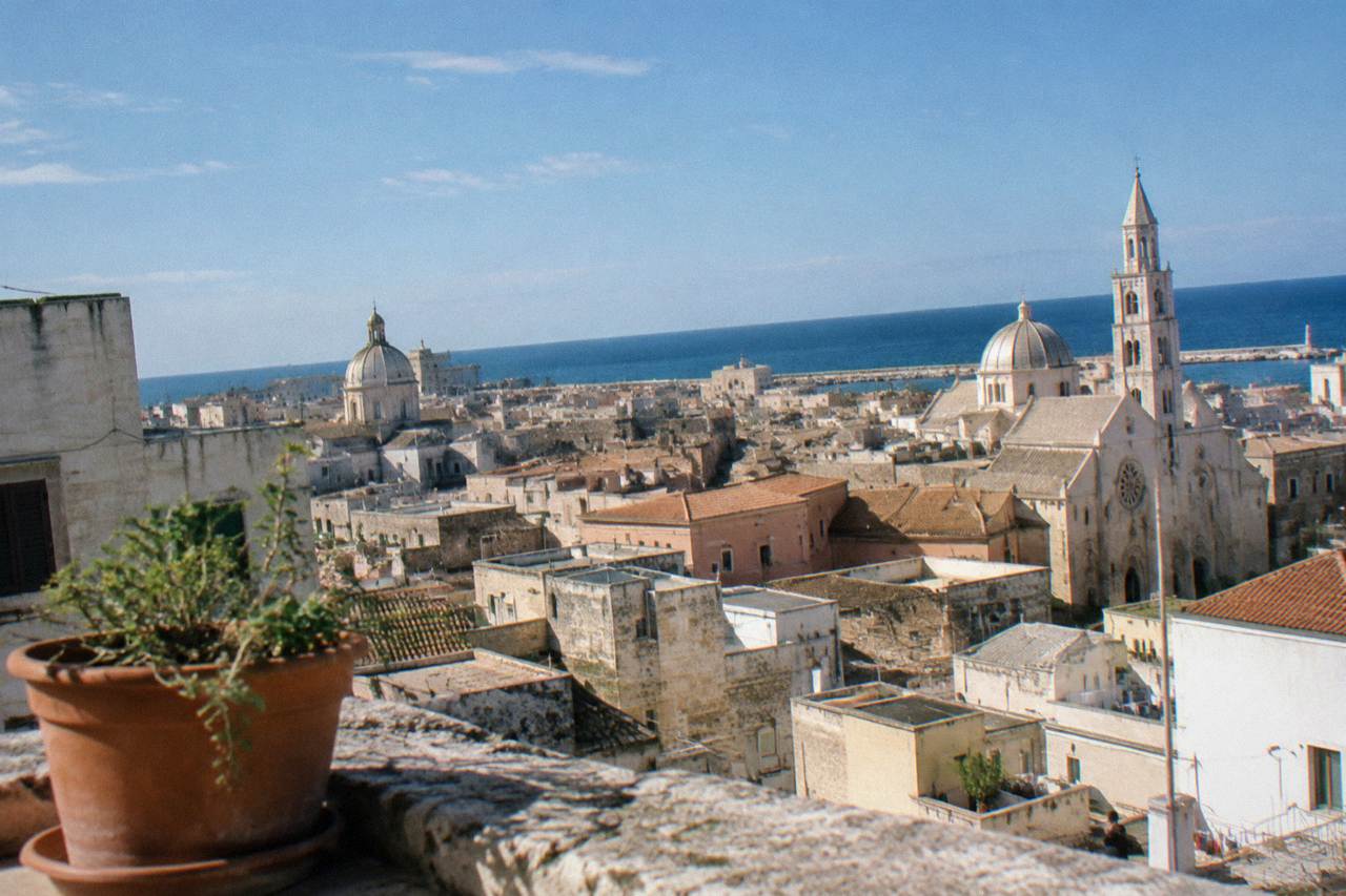 Terracotta and limestone rooftops and church domes of a dense old town seen from a high terrace with the sea beyond