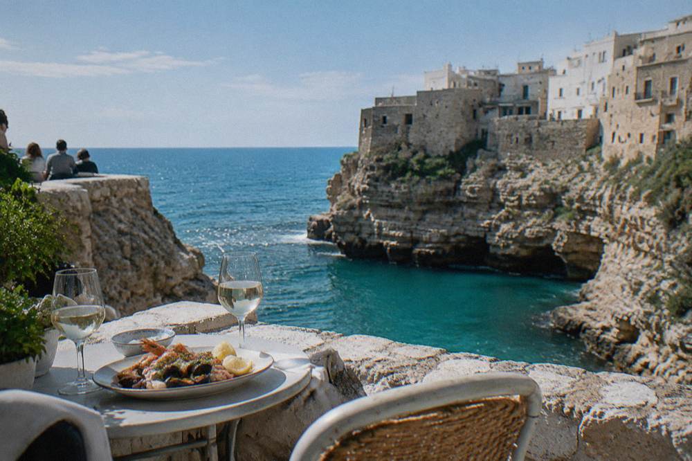 Small round tables on a cliff-edge terrace with glasses and a plate of seafood above blue water