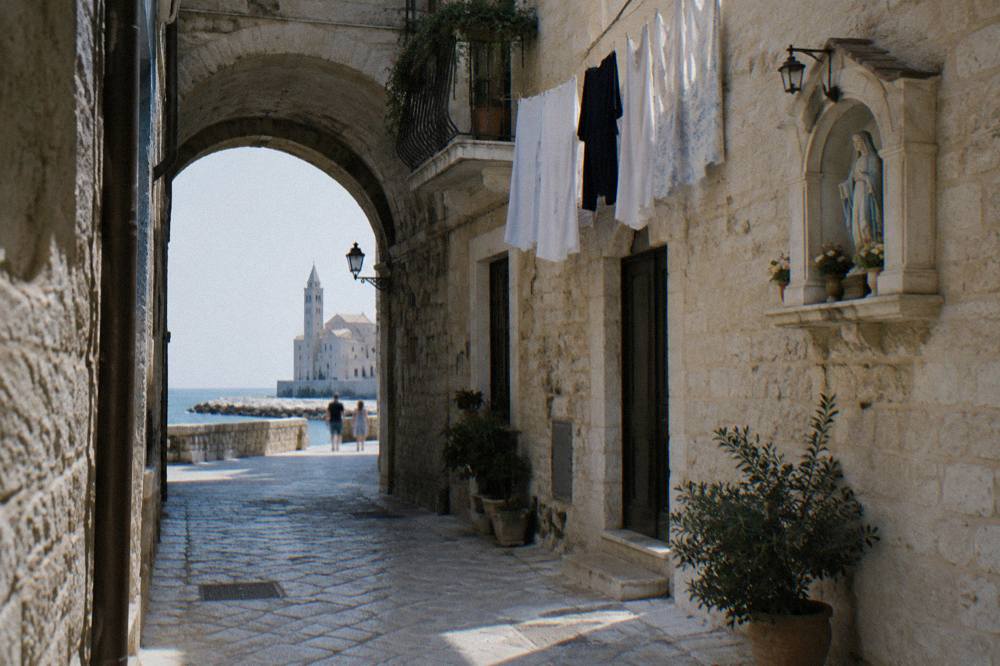 Narrow pale limestone lane with an arch, hanging laundry and a small shrine niche in soft morning light