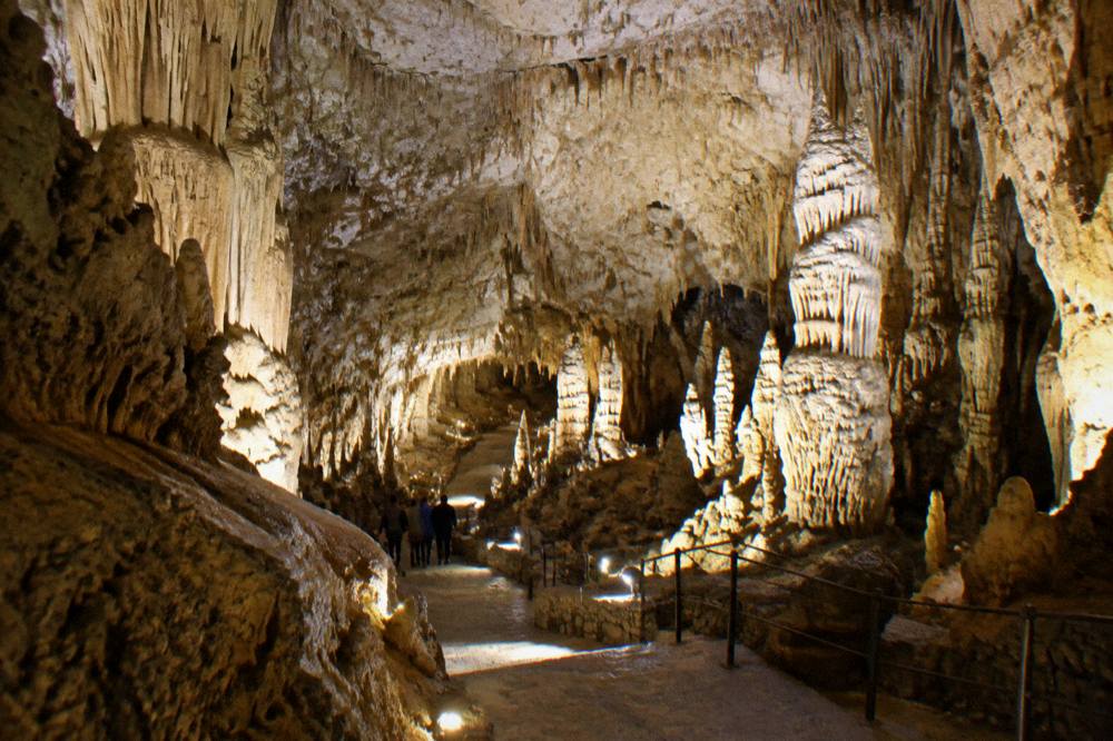 Vast underground cave hall with pale stalactites and stalagmites under warm lights and tiny visitors on a stepped path