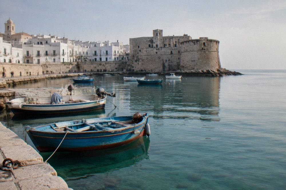 Old harbour of small painted wooden boats with a stone castle on the headland and a whitewashed old town behind