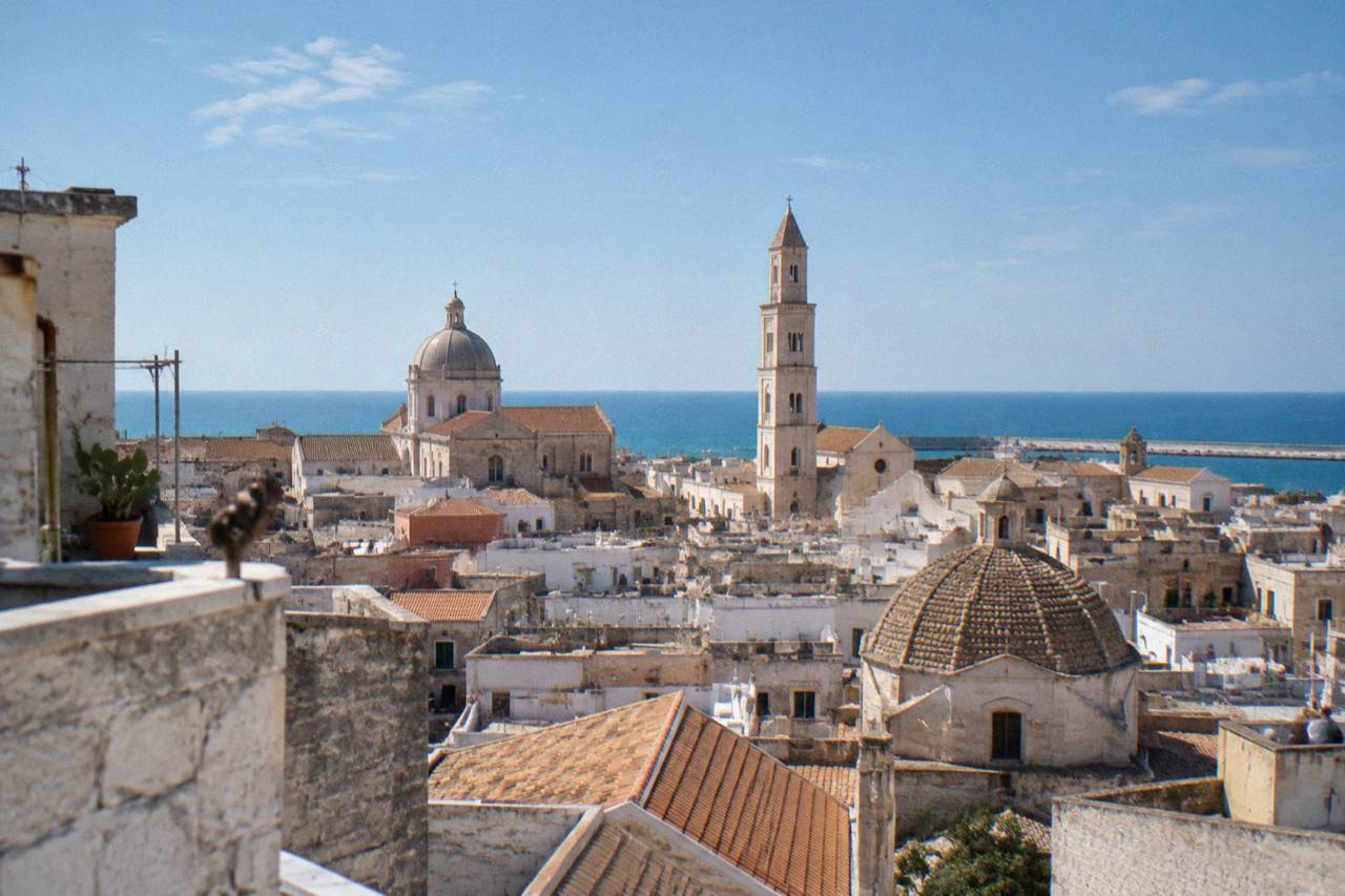 Terracotta and limestone rooftops and church domes of a dense old town seen from a high terrace with the sea beyond