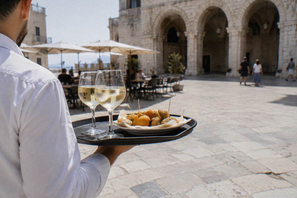 Waiter's tray with two glasses of white wine and fried snacks carried across a pale stone square