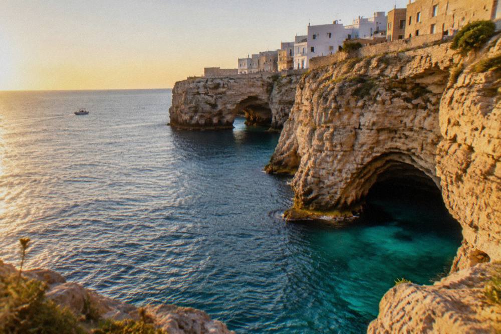 Sea cave openings at the base of tall limestone cliffs with turquoise water and a small boat far off