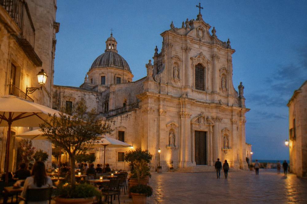 Baroque cathedral facade in warm pale stone on a small square with a dome behind and cafe tables