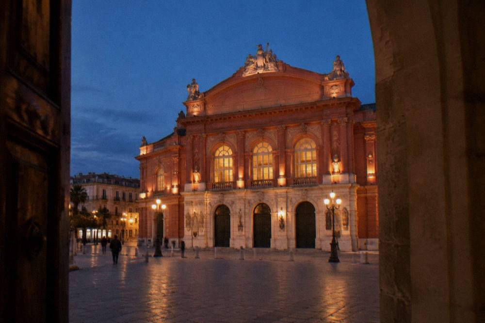Neoclassical theatre facade floodlit at evening with glowing arched windows and a deep blue sky