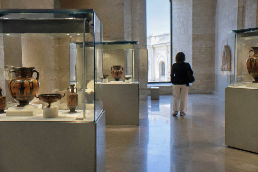 Quiet museum room with glass cases of ancient painted pottery under soft spotlighting and one visitor from behind