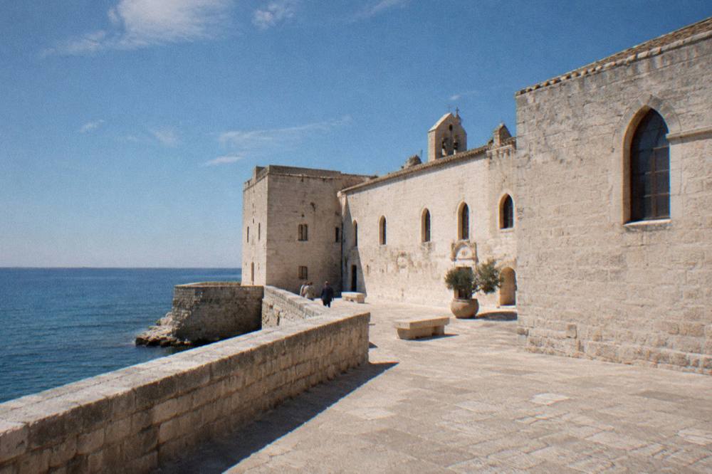 Low pale stone former monastery beside the sea with arched windows and a bare stone terrace
