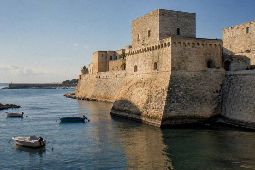 Seaward wall of a pale stone castle beside harbour water with a few small boats in afternoon sun